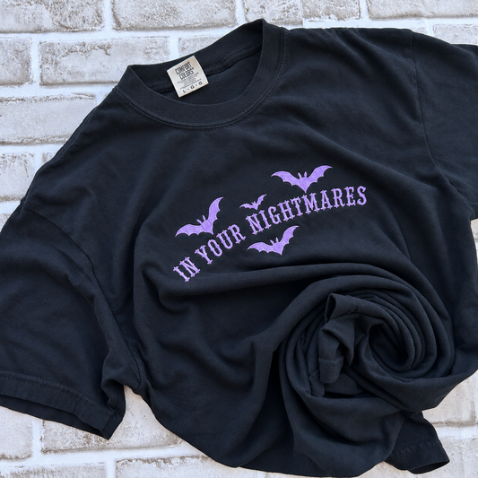 black Comfort Colors T-shirt featuring lavender embroidered gothic text reading “In Your Nightmares,” surrounded by four embroidered bats, styled on a white background.