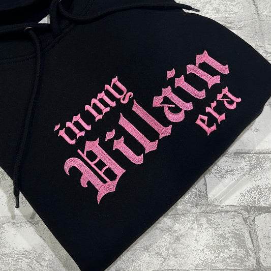 A black hoodie with the phrase 'In My Villain Era' embroidered in pink on the front.