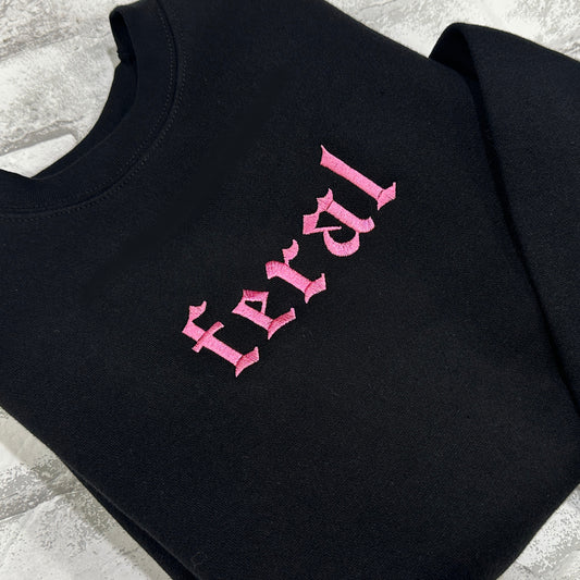 A black crewneck sweatshirt with the word 'feral' embroidered in pink on the front.