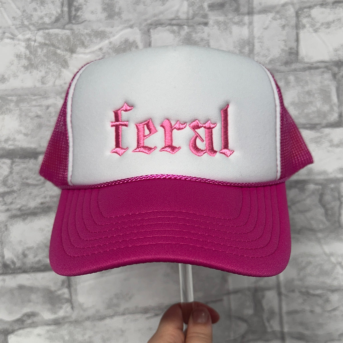 Front view of a pink and white mesh trucker hat with pink embroidered text reading “feral.”