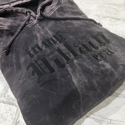 Black mineral-wash pullover hoodie folded flat, featuring black gothic lettering that reads ‘in my Villain era.