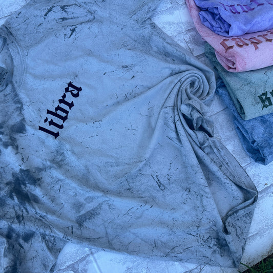 A collection of embroidered t-shirts with various zodiac signs displayed on a stone surface. The main shirt is gray with the zodiac Libra embroidered on the front, and others have different signs like Aries, Capricorn, Scorpio, and Pisces.