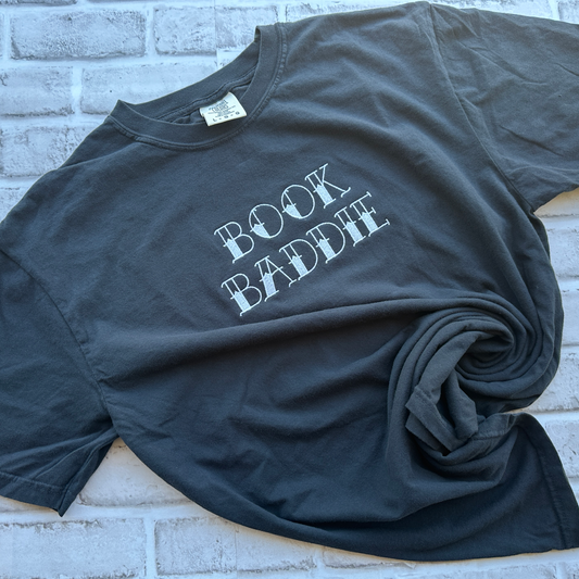 A black short-sleeve t-shirt with the phrase 'BOOK BADDIE' embroidered in a distressed white font across the chest, laid flat against a white background.
