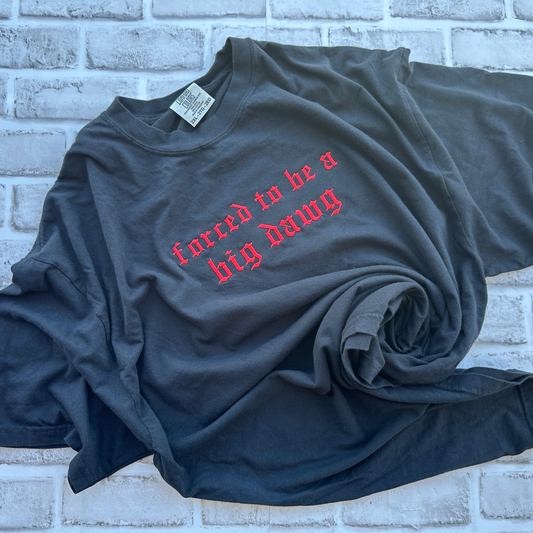Black Comfort Colors T-shirt featuring red Old English embroidered text across the chest that reads “forced to be a big dawg,” shown flat on a white background.