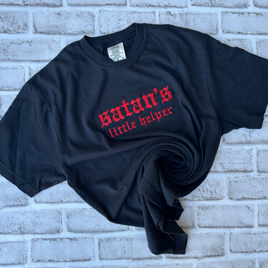 Black Comfort Colors short-sleeve T-shirt featuring red Old English gothic embroidery reading “satan’s little helper” across the upper chest. “Satan’s” appears in larger lettering above the smaller “little helper” text. Classic crewneck collar with a relaxed fit, displayed flat against a white background.