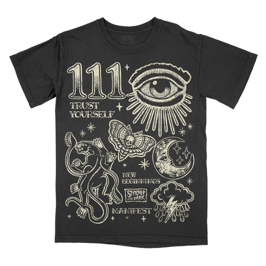 black T-shirt featuring a distressed 111 angel number design with an all-seeing eye, black panther, moth, moon, stars, and storm cloud, with text reading “Trust Yourself,” “New Beginnings,” and “Manifest,” plus Stitched in the Dark branding.