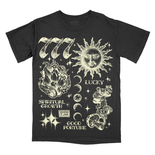 black T-shirt featuring a distressed 777 angel number design with a celestial sun, crystals, moon phases, mushrooms, and stars, with text reading “Lucky,” “Spiritual Growth,” and “Good Fortune,” plus Stitched in the Dark branding.