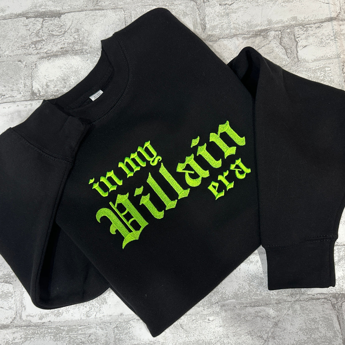 Black crewneck sweatshirt with green embroidered text 'in my Villain era' on the front.