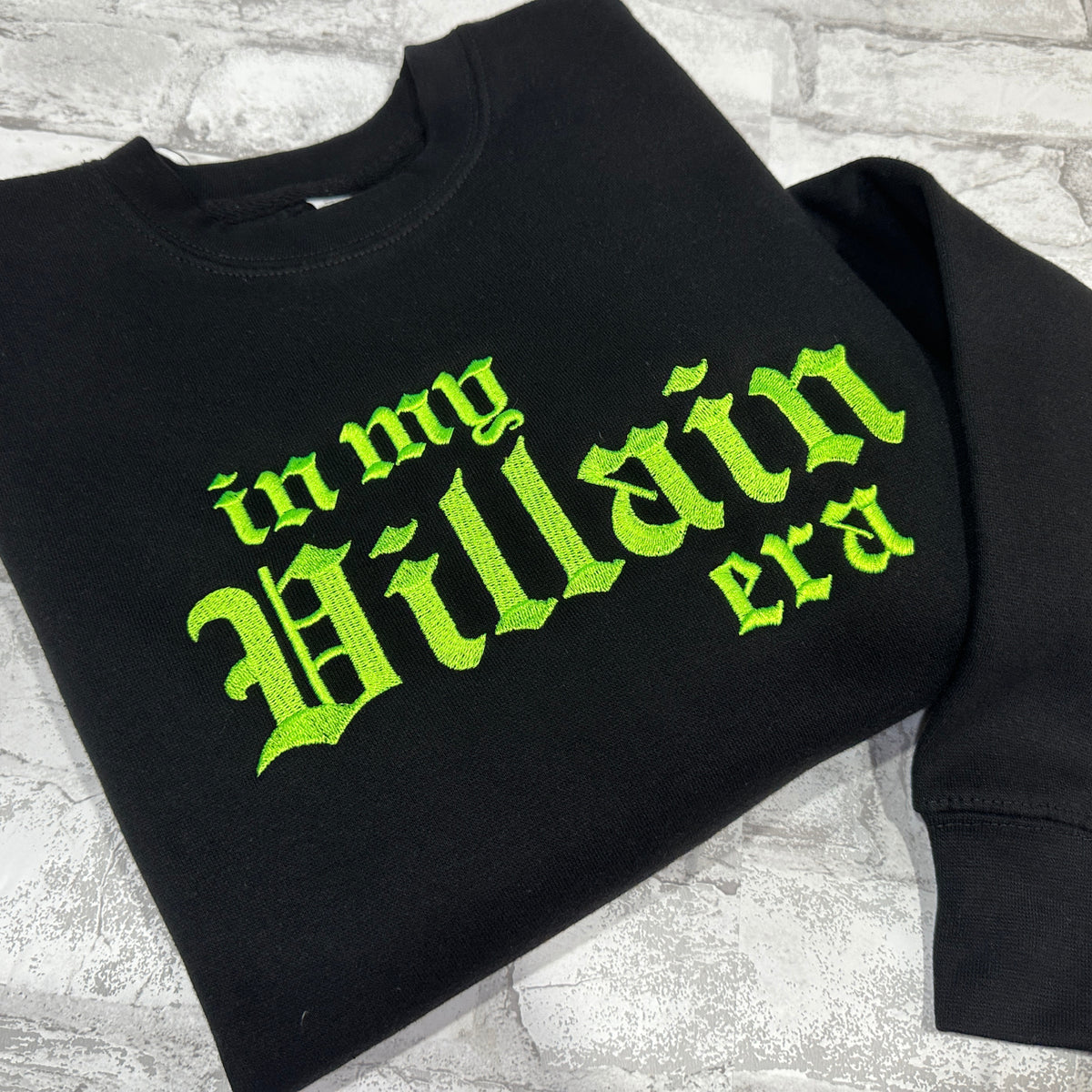 Black crewneck sweatshirt with green embroidered text 'in my Villain era' on the front.