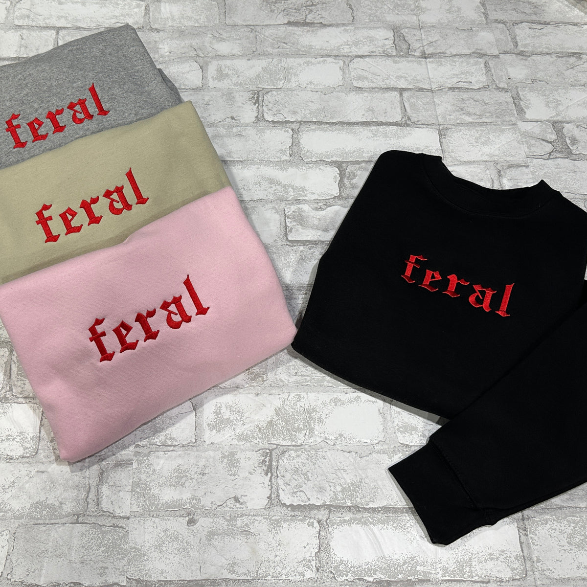Four crewneck sweatshirts in grey, tan, pink, and black colors, each with the word 'feral' embroidered in red on the front.