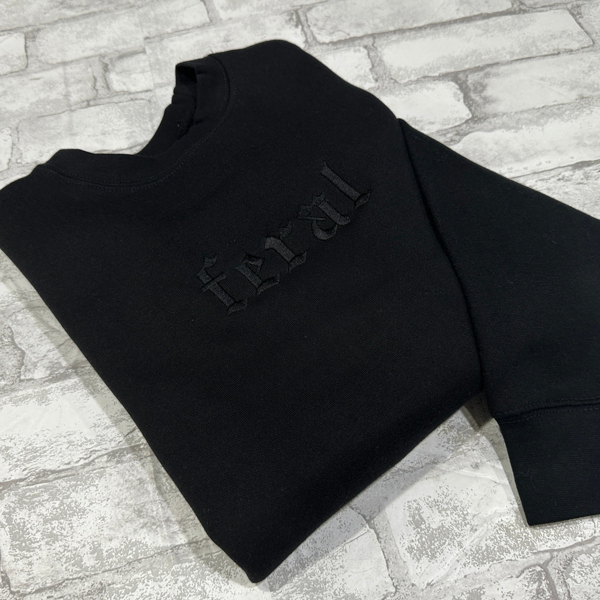 Black sweatshirt with black embroidered gothic text reading “feral” on the chest.