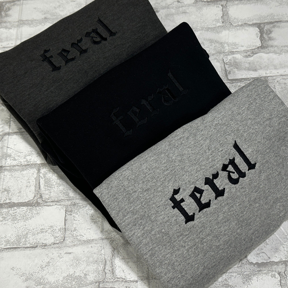 Three crewneck sweatshirts, one in black, one in dark gray,  and one in gray, with the word 'feral' embroidered in bold lettering across the front.