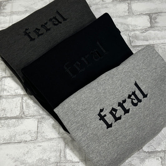 Three crewneck sweatshirts, one in black, one in dark gray,  and one in gray, with the word 'feral' embroidered in bold lettering across the front.