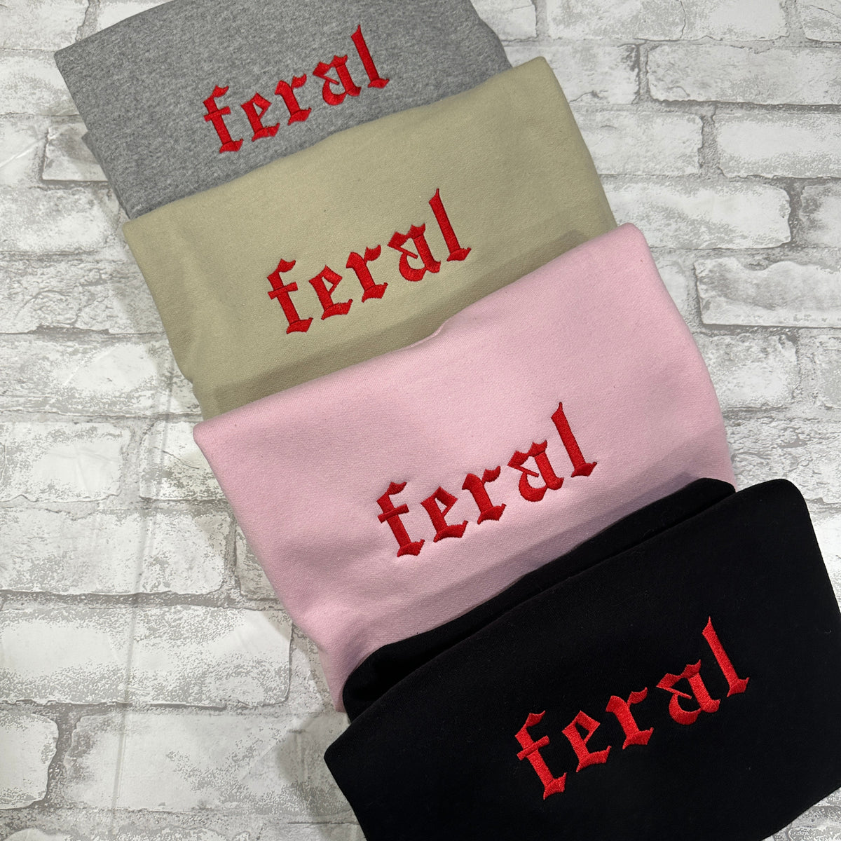 Four crewneck sweatshirts in grey, tan, pink, and black colors, each with the word 'feral' embroidered in red on the front.