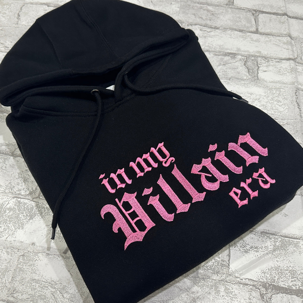 A black hoodie with the phrase 'In My Villain Era' embroidered in pink on the front.
