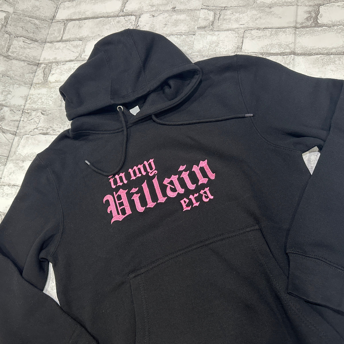 A black hoodie with the phrase 'In My Villain Era' embroidered in pink on the front.