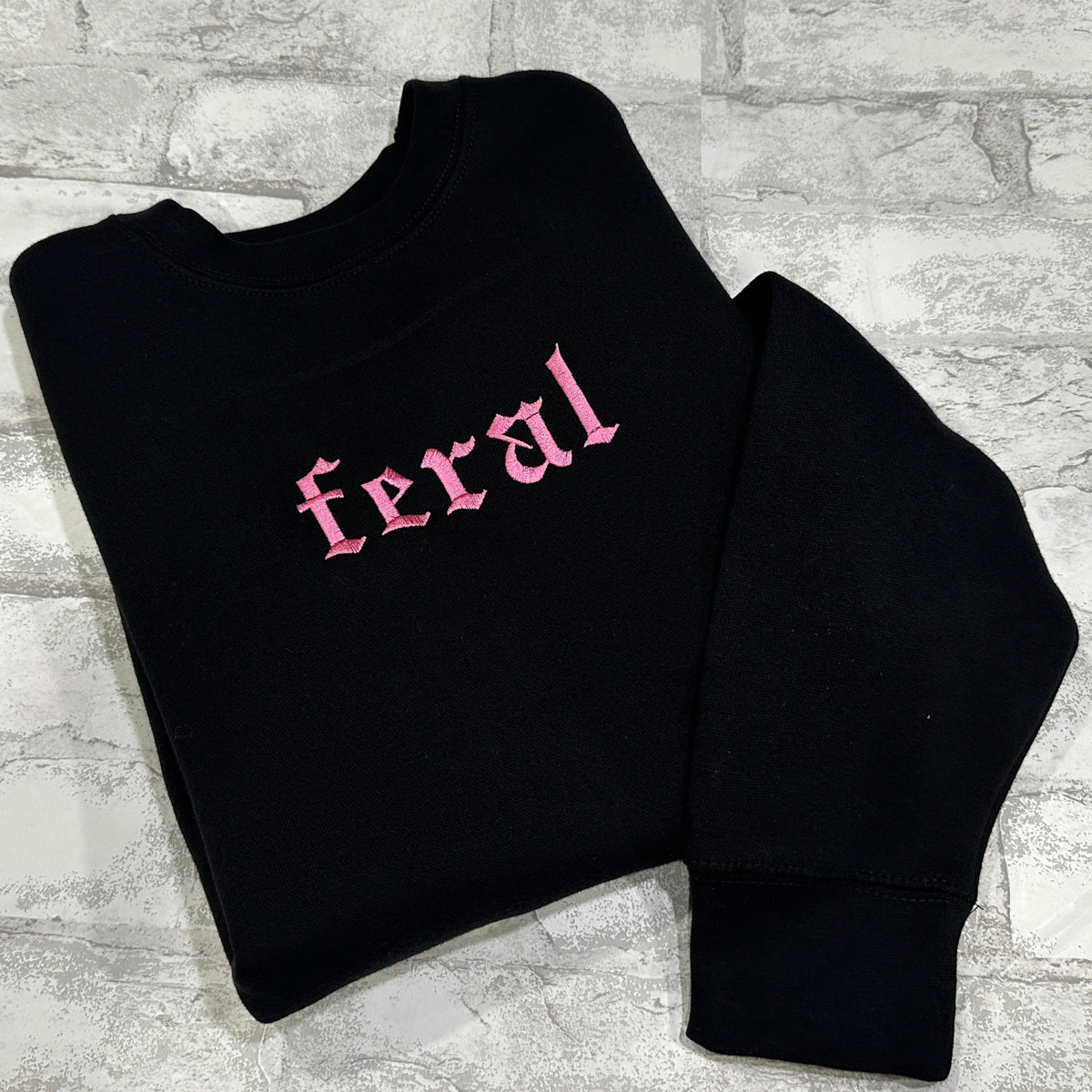 A black crewneck sweatshirt with the word 'feral' embroidered in pink on the front.