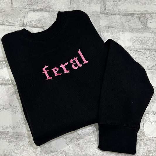 A black crewneck sweatshirt with the word 'feral' embroidered in pink on the front.