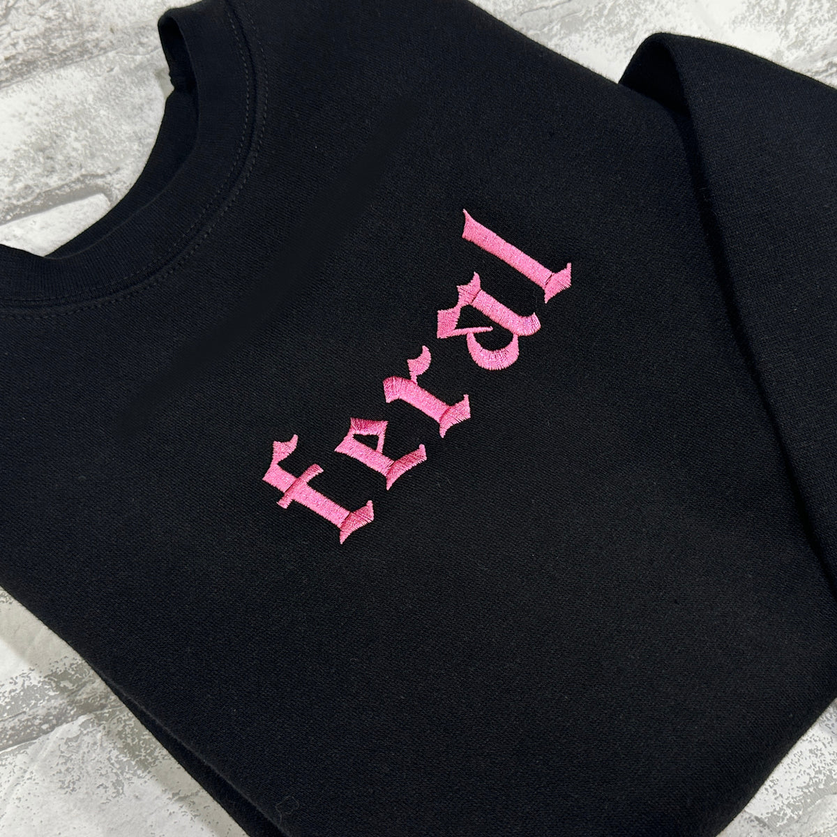 A black crewneck sweatshirt with the word 'feral' embroidered in pink on the front.