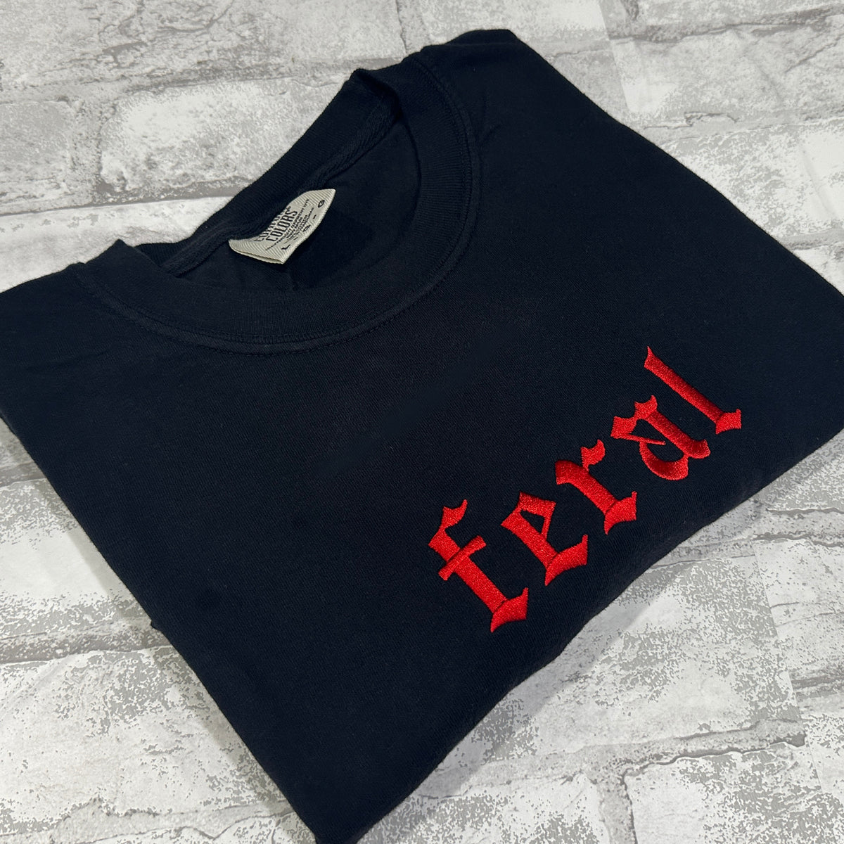 A black Comfort Colors embroidered tee with the word 'feral' in red lettering on the front.