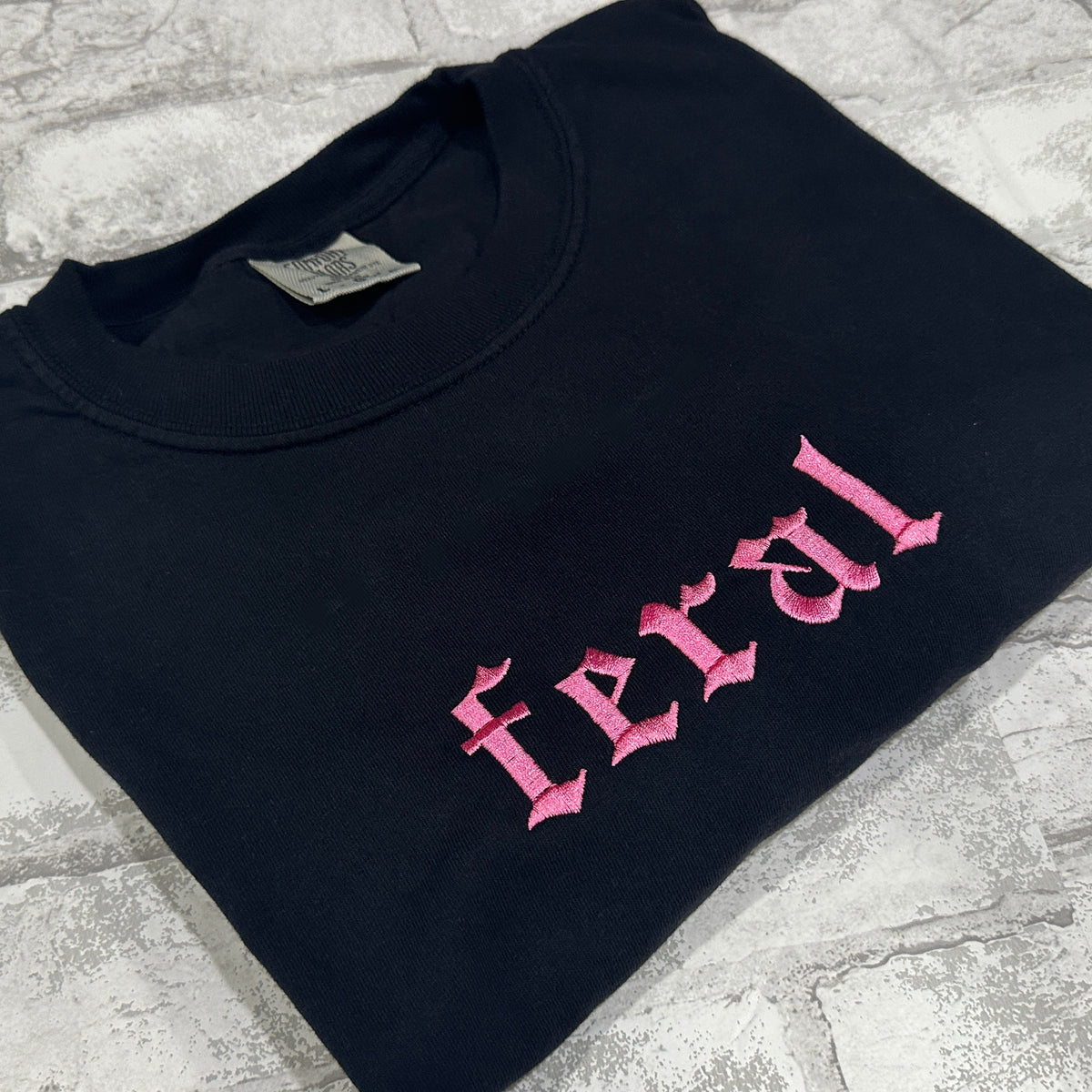 Black Comfort color shirt with pink embroidered gothic text reading “feral” on the chest.