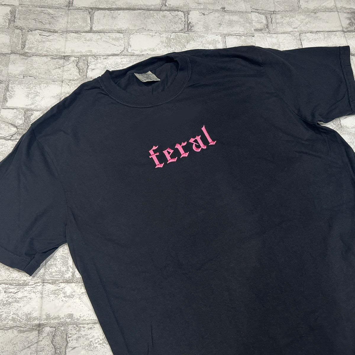 Black Comfort color shirt with pink embroidered gothic text reading “feral” on the che