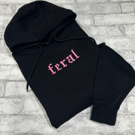 A black hoodie with the word 'feral' embroidered in pink on the front.