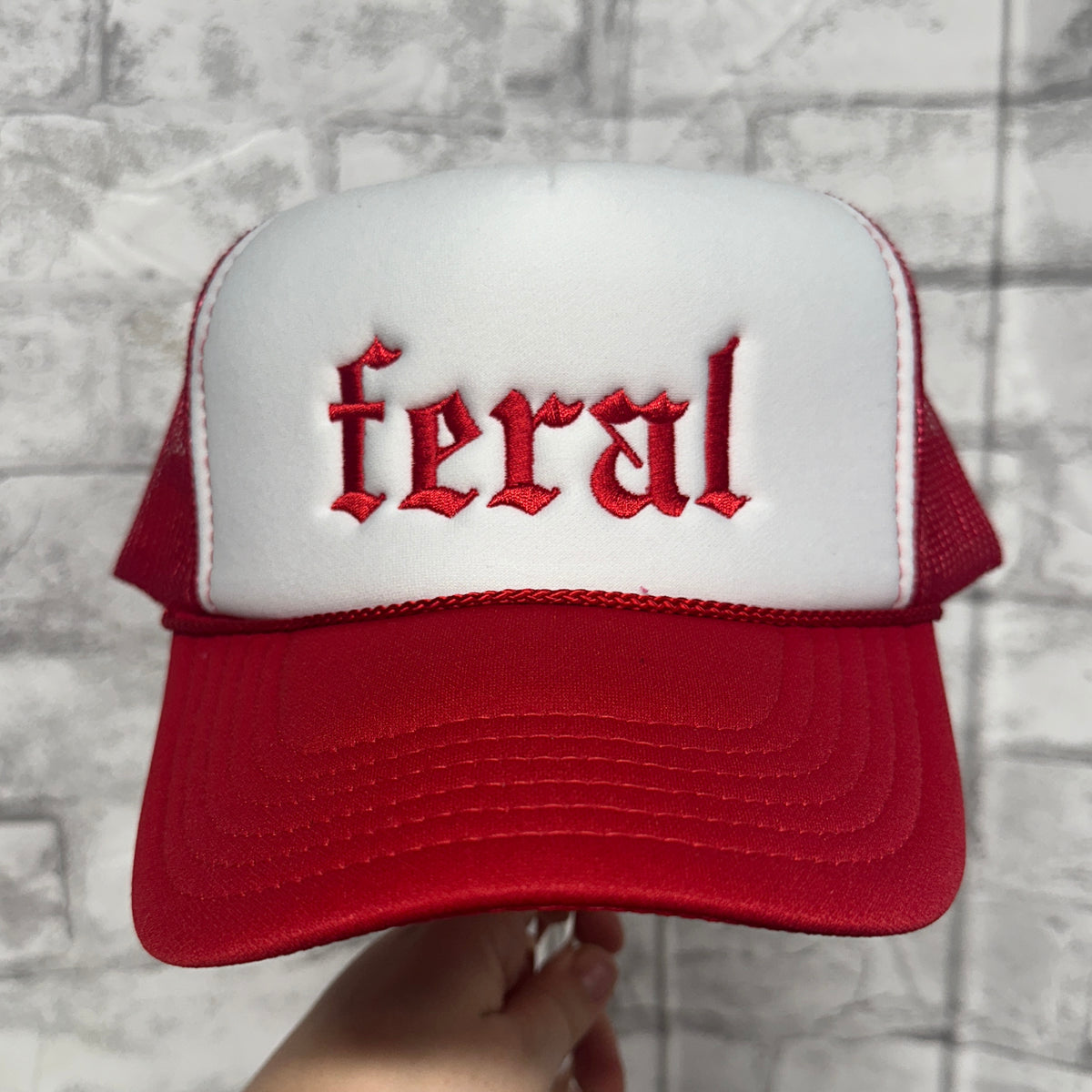 Front view of a red green and white mesh trucker hat with red embroidered text reading “feral.”