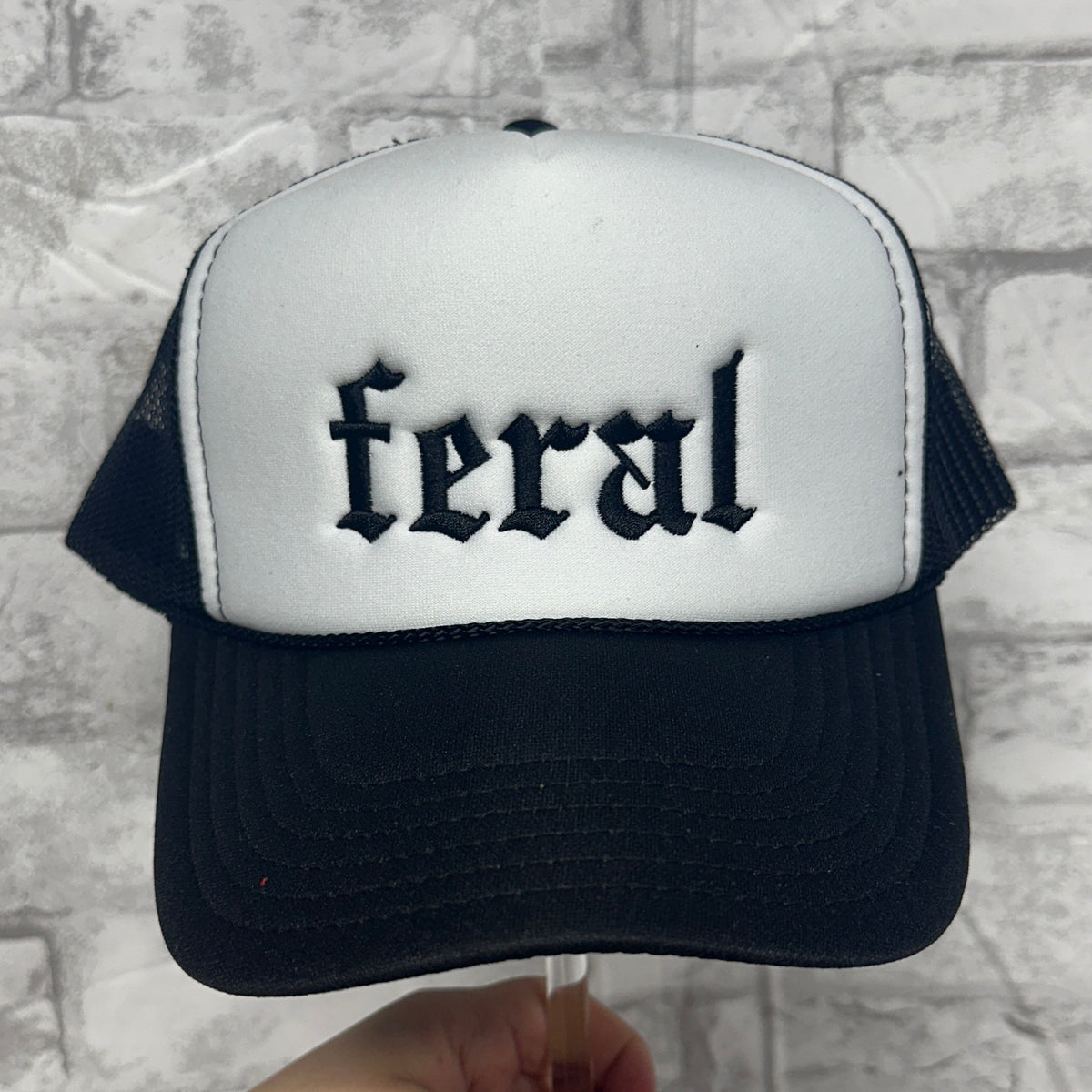 Front view of a black and white mesh trucker hat with black embroidered text reading “feral.”