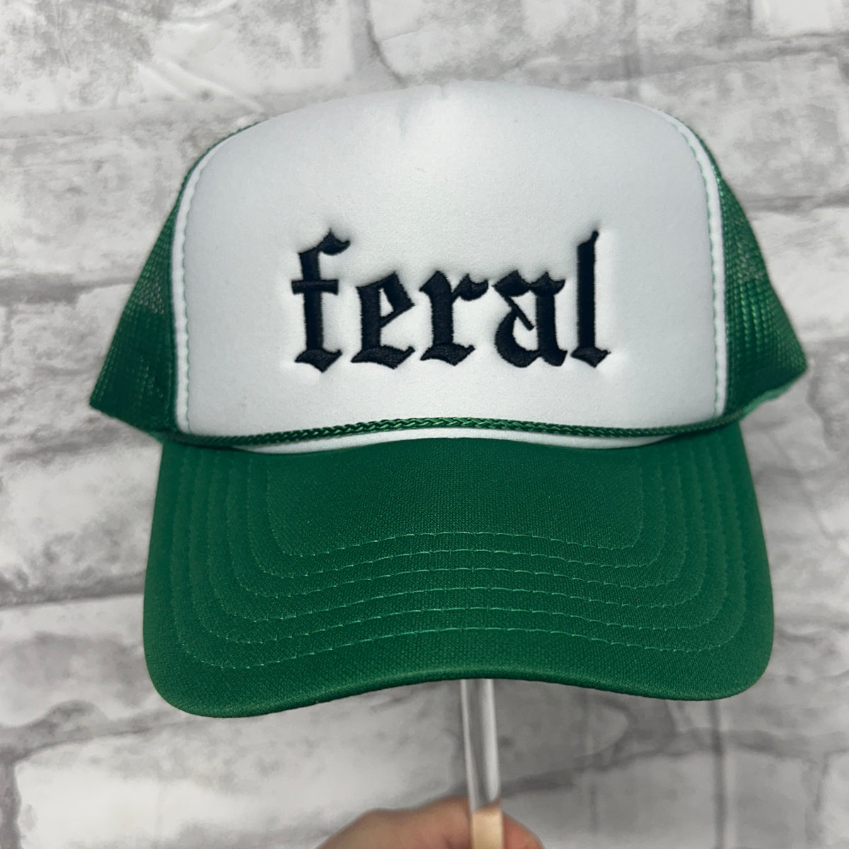 Front view of a  green and white mesh trucker hat with green embroidered text reading “feral.”