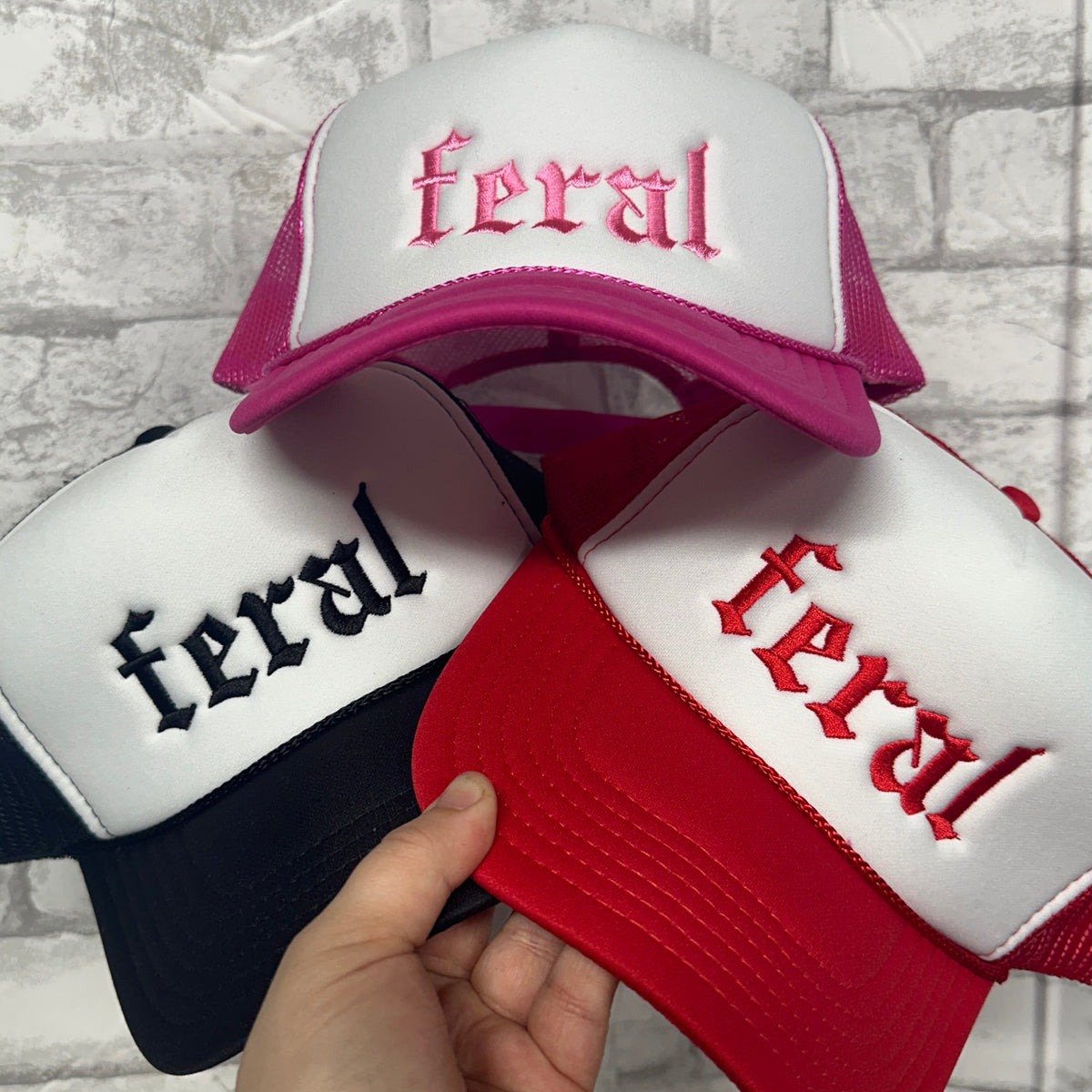 Three embroidered foam trucker hats in red, pink, and black, all with a white panel and each with the word 'Feral' stitched on the front.