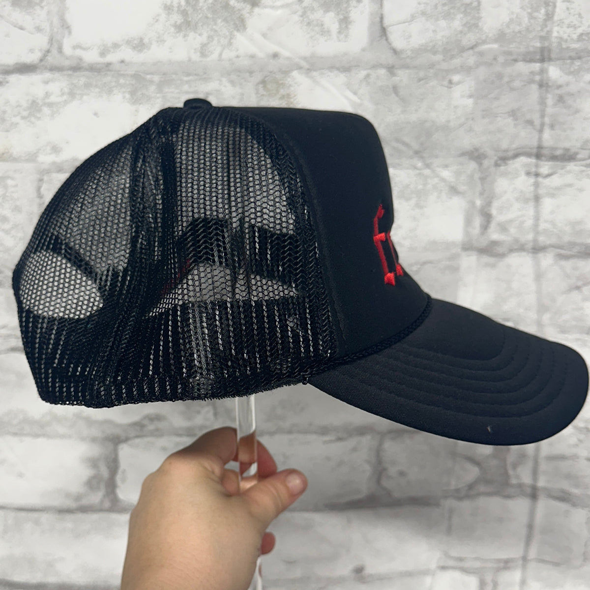 Side view of a black mesh trucker hat with red embroidered text on the front panel.