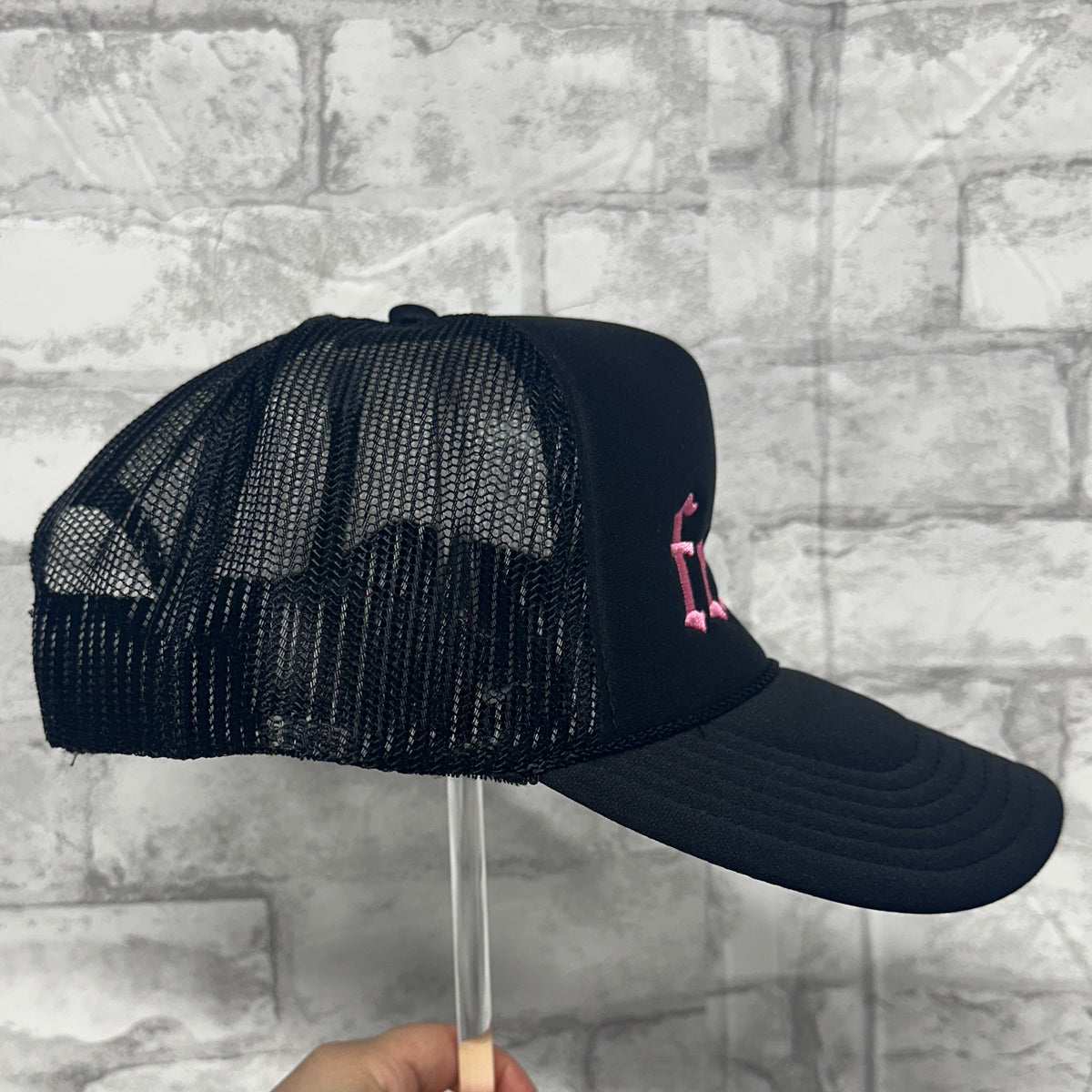 Side view of a black mesh trucker hat with pink embroidered text on the front panel.