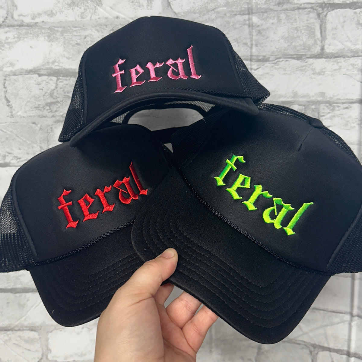 Three black trucker hats with embroidered text 'Feral' in red, green, and pink, on a grey background.
