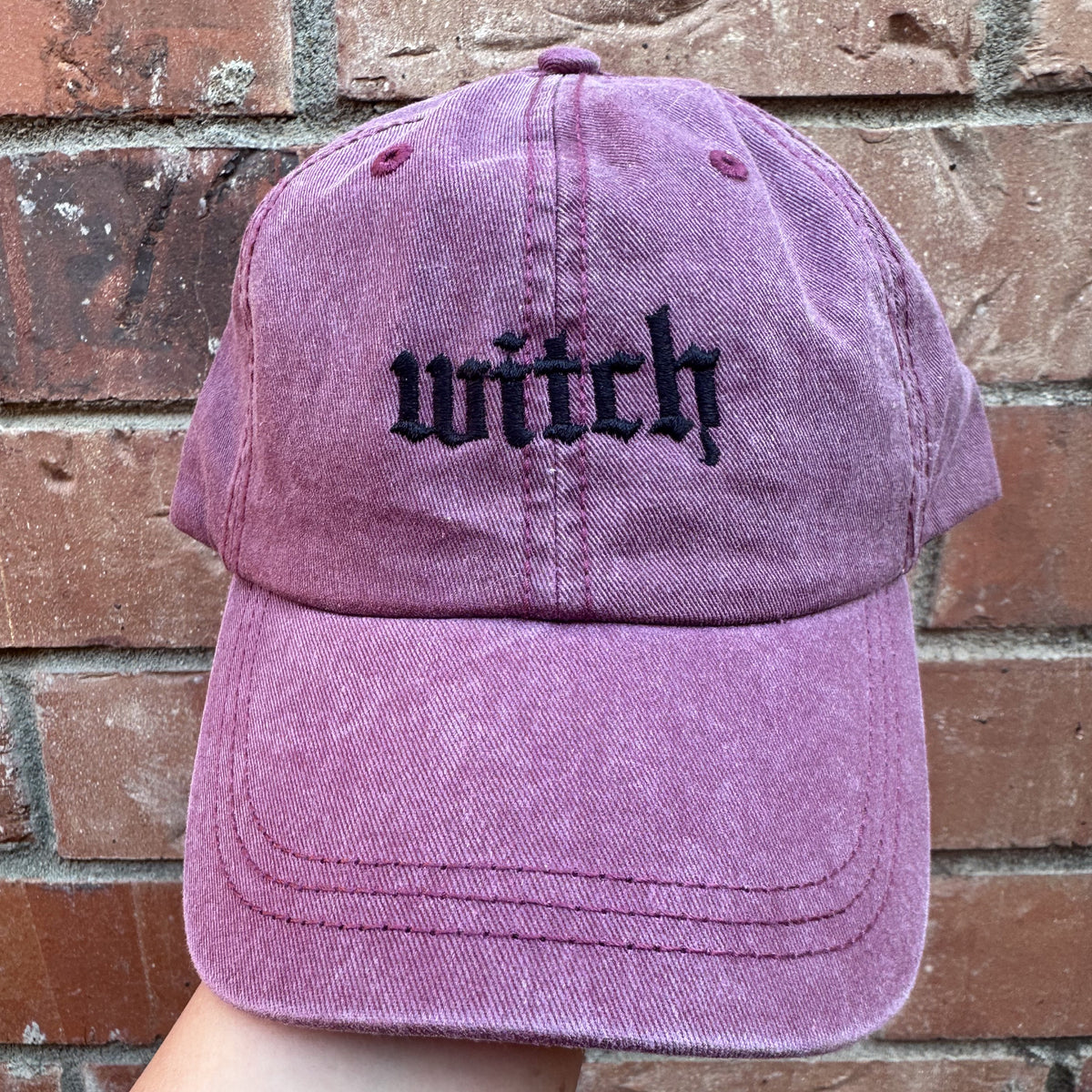 A pigment dyed baseball cap in a purple hue with the word 'witch' embroidered in black thread on the front.