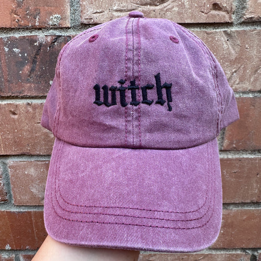 A pigment dyed baseball cap in a purple hue with the word 'witch' embroidered in black thread on the front.