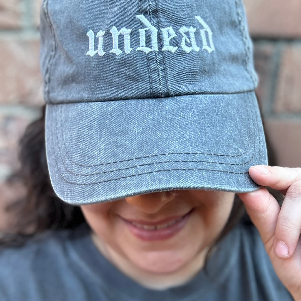 Pigment dyed black  baseball cap with curved brim, featuring white embroidered text reading “undead” on the front panel being modeled on girl. 