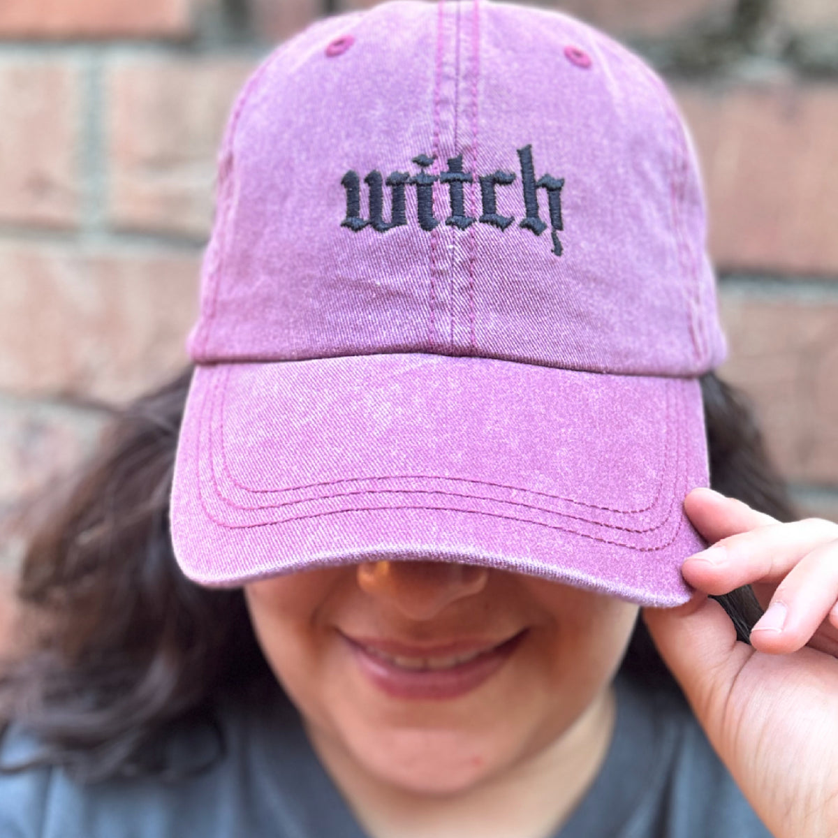 Pigment dyed purple baseball cap with curved brim, featuring black embroidered text reading “witch” on the front panel model by female. 