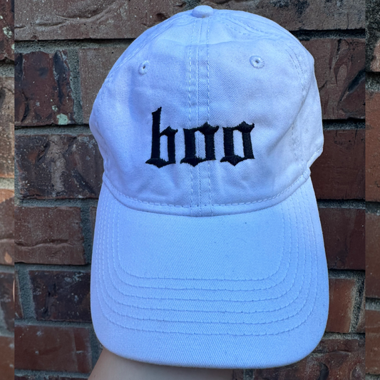Pigment dyed white baseball cap with curved brim, featuring black embroidered text reading “boo” on the front panel.