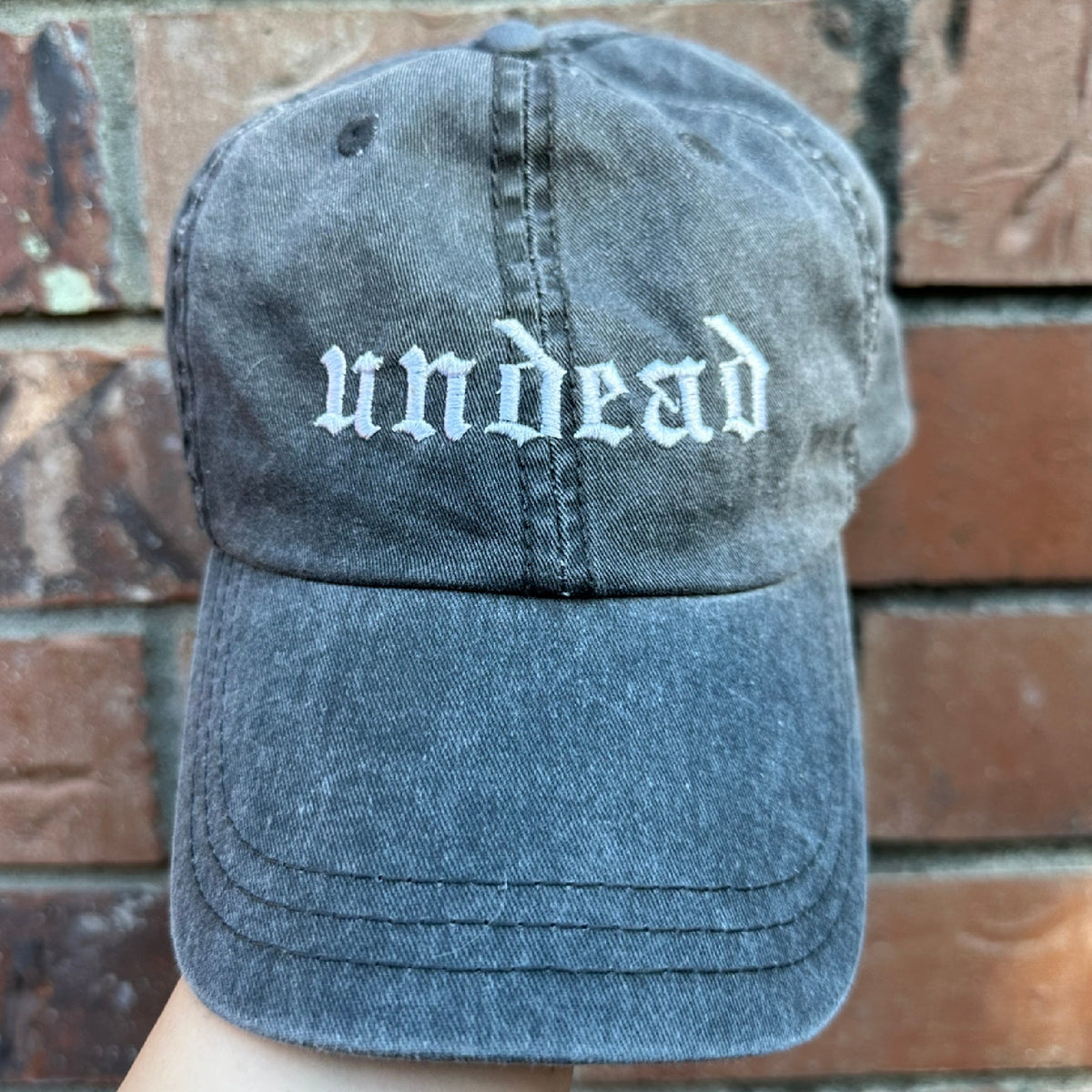 Pigment dyed black  baseball cap with curved brim, featuring white embroidered text reading “undead” on the front panel.