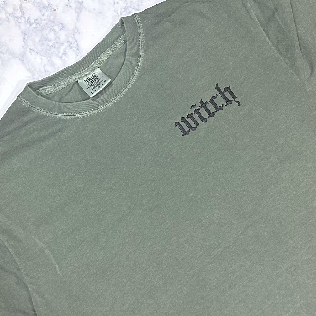 Green Moss Comfort Colors t-shirt with the word 'witch' embroidered on the left chest area in black thread.