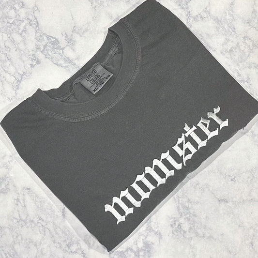 A pepper Comfort Colors t-shirt with the text 'momster' embroidered in white thread on the front.