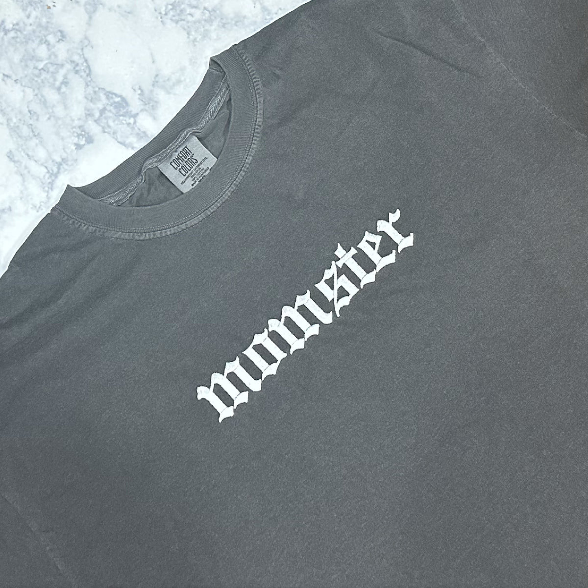 A pepper Comfort Colors t-shirt with the text 'momster' embroidered in white thread on the front.