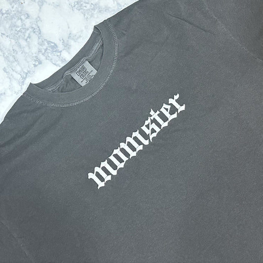 A pepper Comfort Colors t-shirt with the text 'momster' embroidered in white thread on the front.