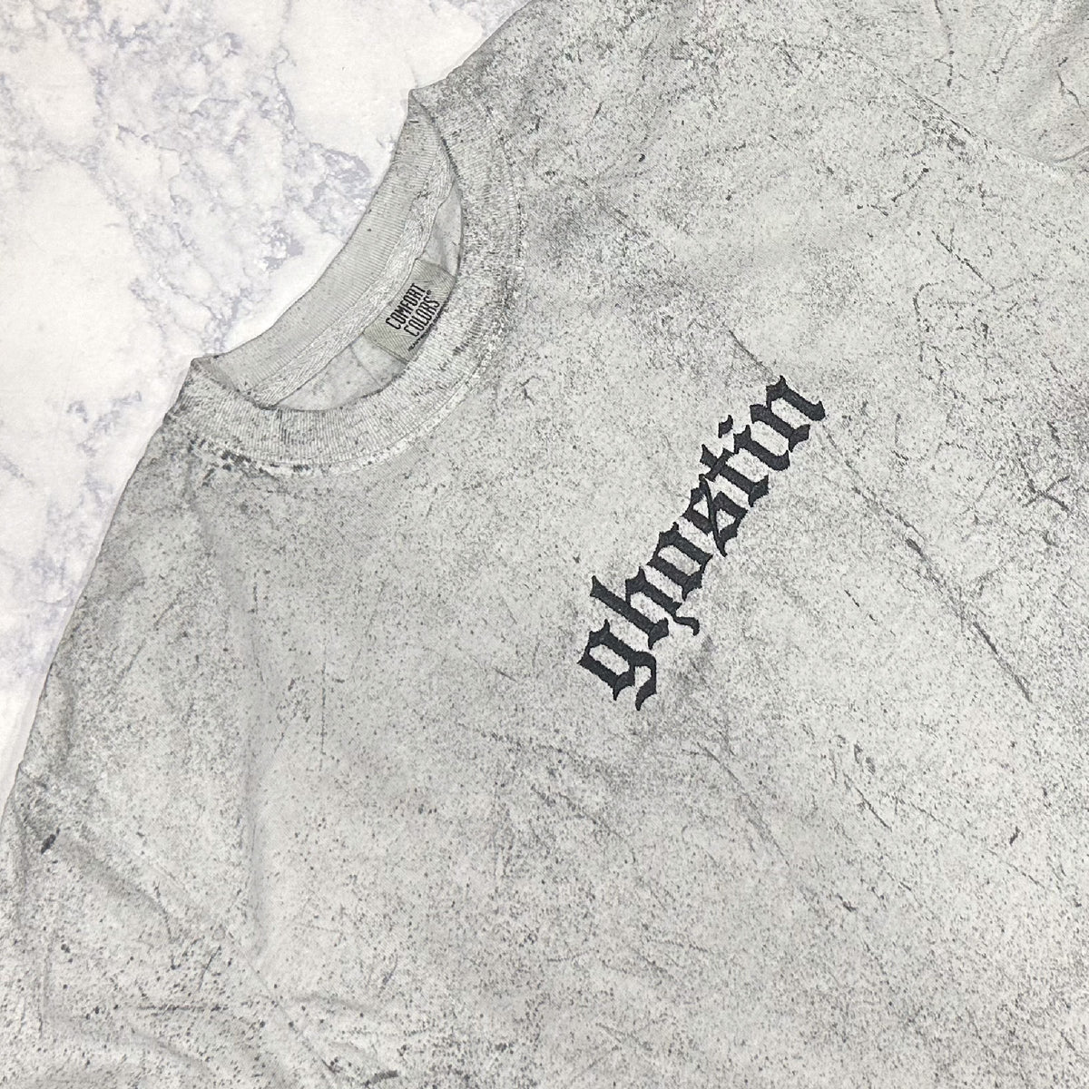 Light gray mineral-washed short-sleeve T-shirt laid flat, featuring small tonal embroidered text reading “ghostin” on the chest.