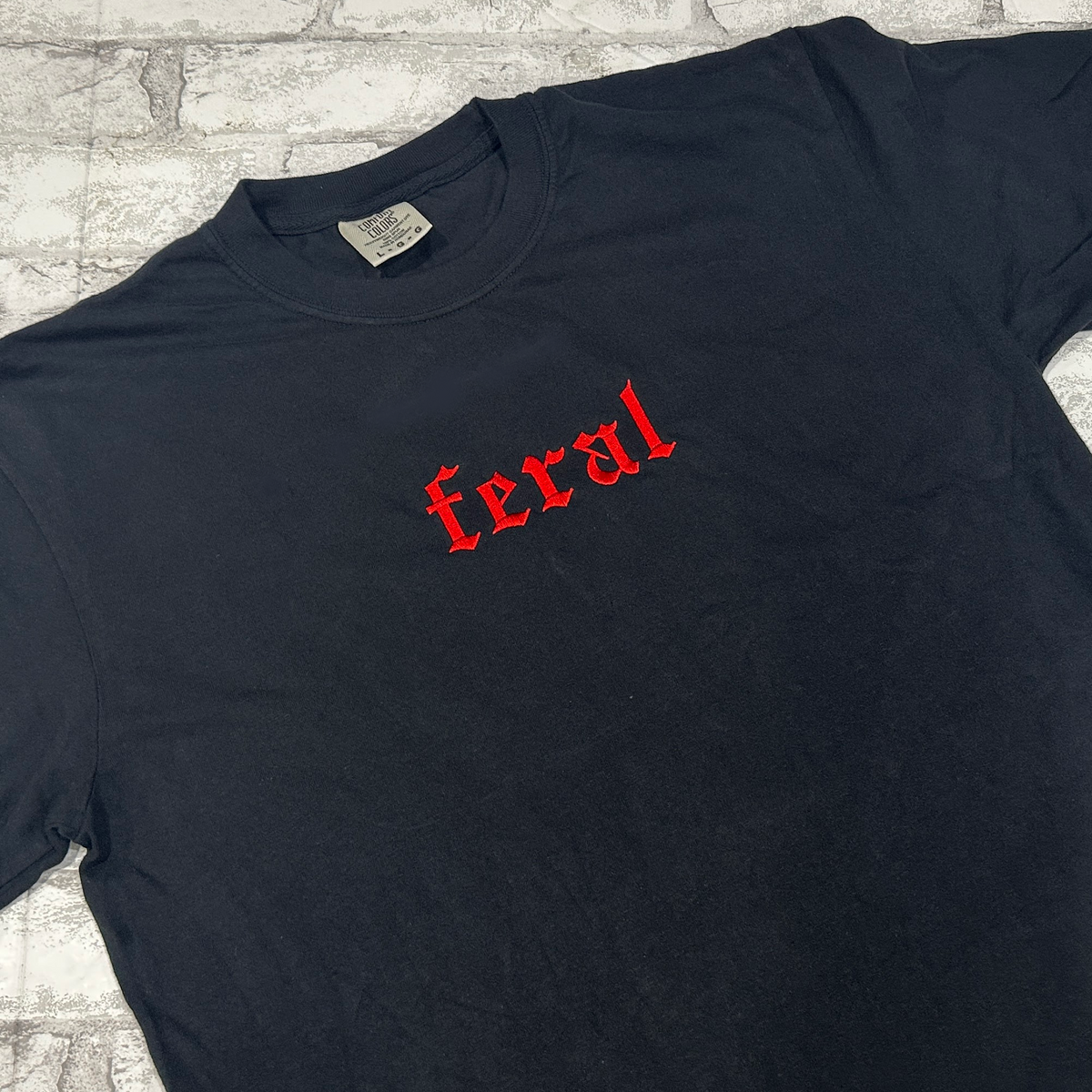 A black Comfort Colors embroidered tee with the word 'feral' in red lettering on the front.