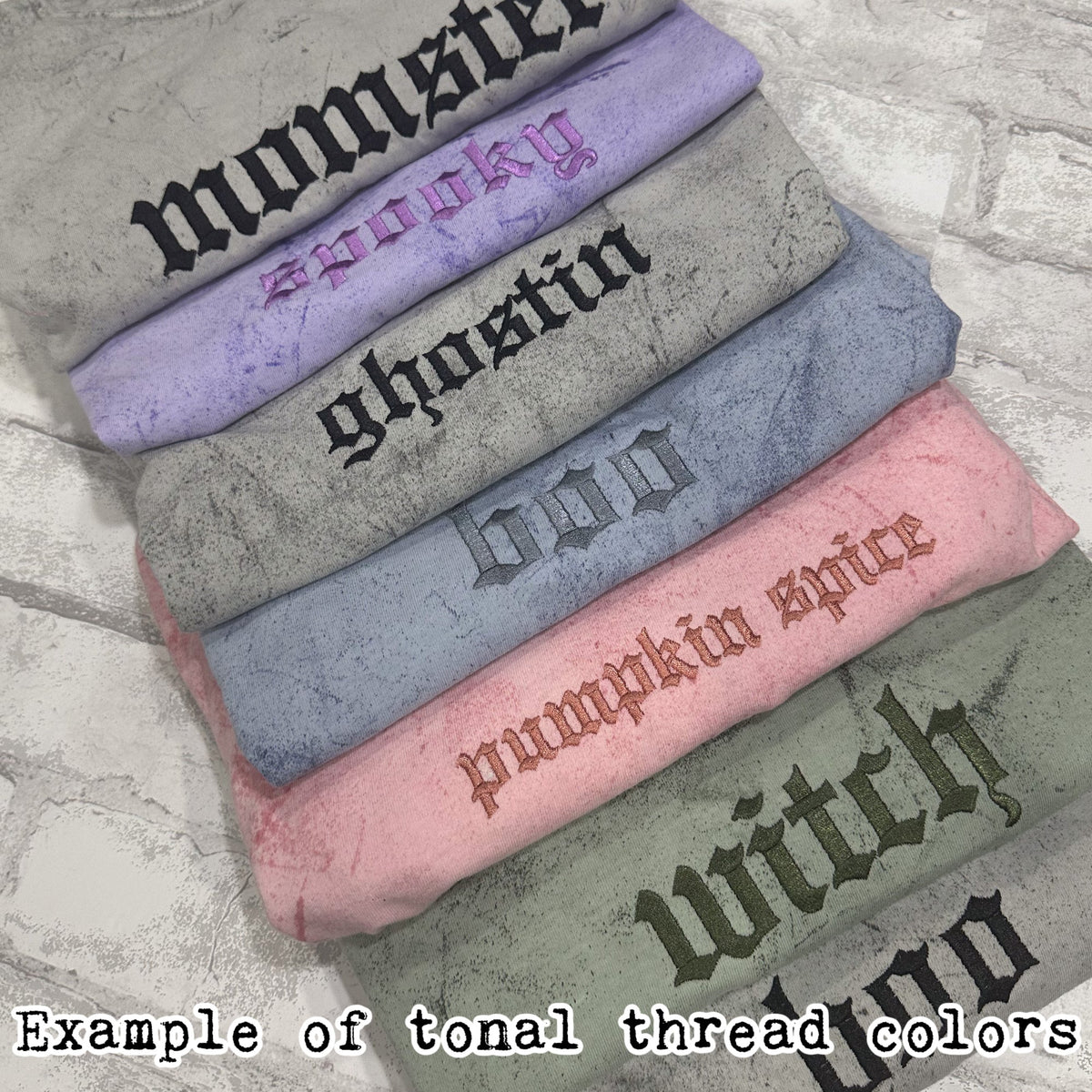 Stack of folded mineral-washed sweatshirts in multiple colors, each with tonal embroidered text showing examples of matching thread colors.