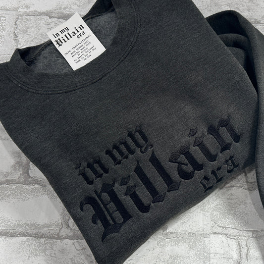 A dark gray embroidered crewneck sweatshirt with the text 'in my Villain Era' displayed on the front.