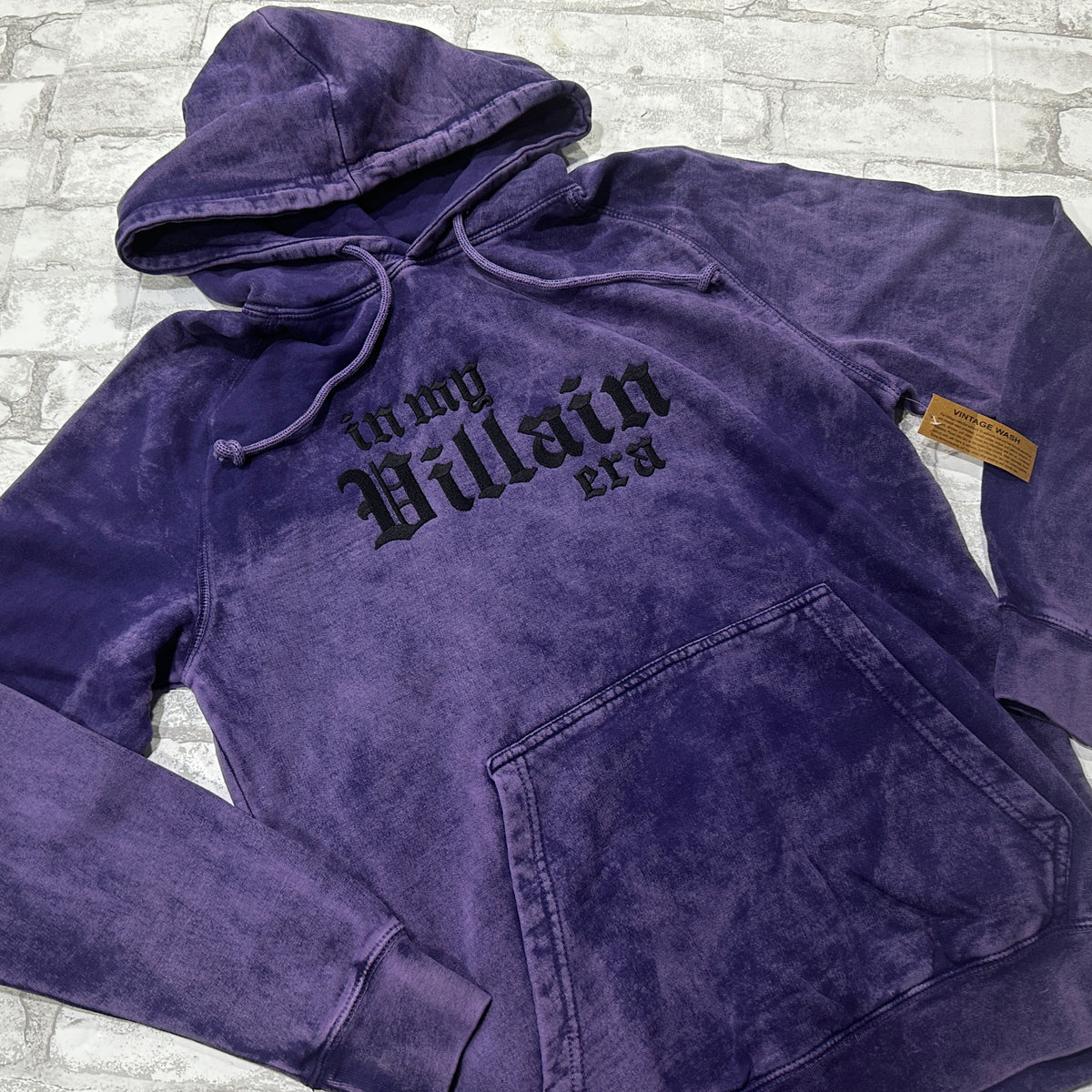 Purple mineral-wash pullover hoodie layed flat, featuring black gothic lettering that reads ‘in my Villain era.