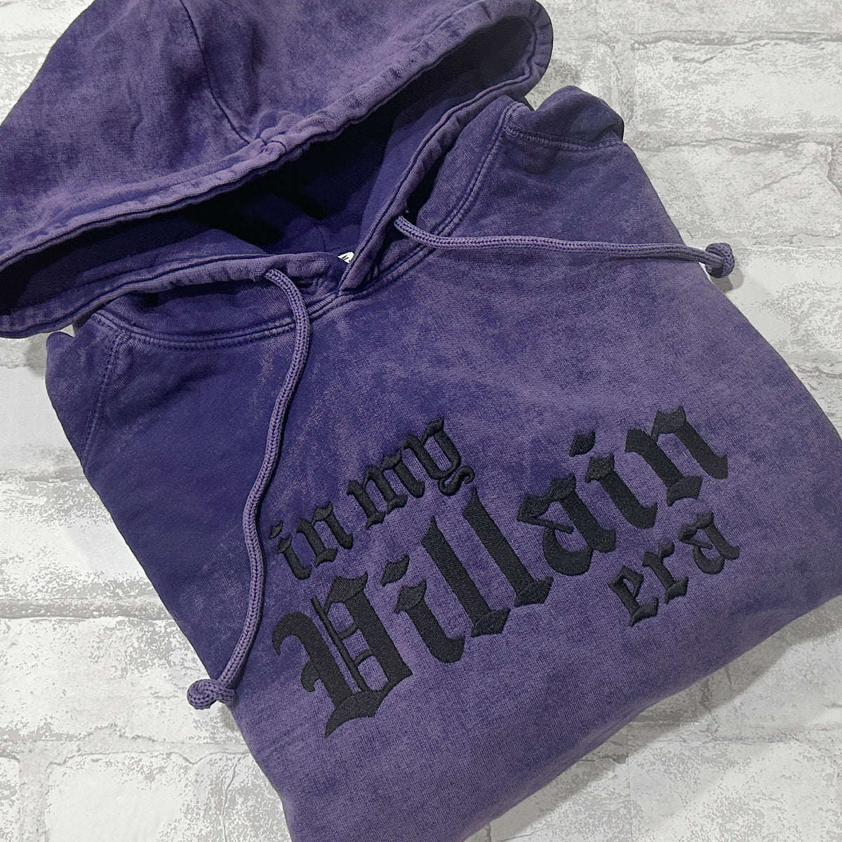 Purple mineral-wash pullover hoodie folded flat, featuring black gothic lettering that reads ‘in my Villain era.